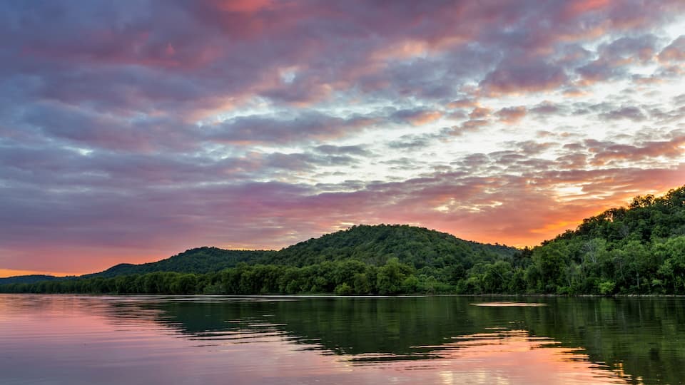 A calm lake with a forested hill in the background and a colorful sky.