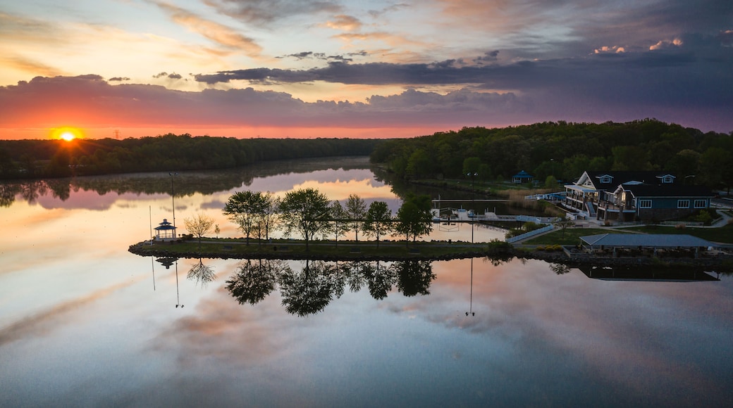 A sunset over a calm lake with a gazebo, dock, and buildings on the shore.