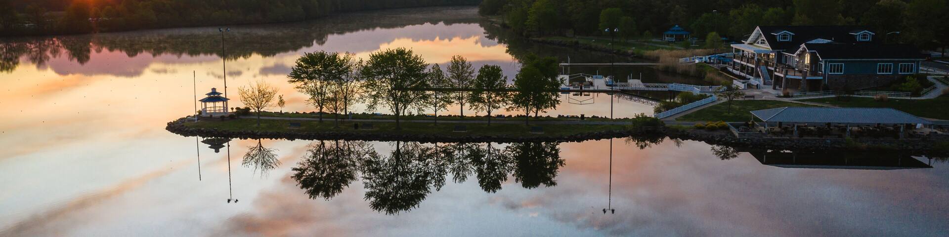 A sunset over a calm lake with a gazebo, dock, and buildings on the shore.