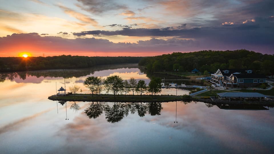 A sunset over a calm lake with a gazebo, dock, and buildings on the shore.