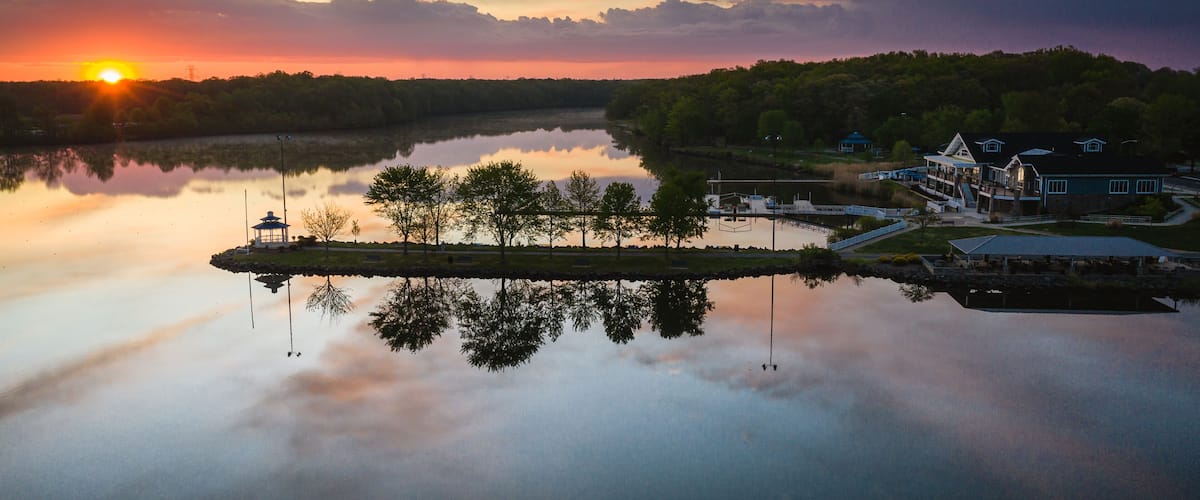 A sunset over a calm lake with a gazebo, dock, and buildings on the shore.