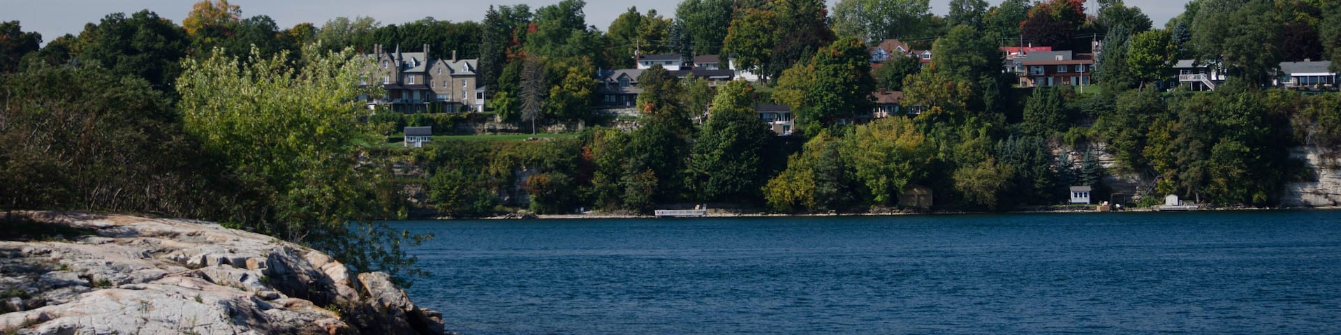 A calm lake with rocky shores, surrounded by dense trees and houses on a hillside.