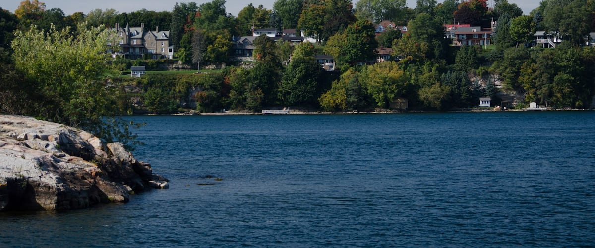 A calm lake with rocky shores, surrounded by dense trees and houses on a hillside.