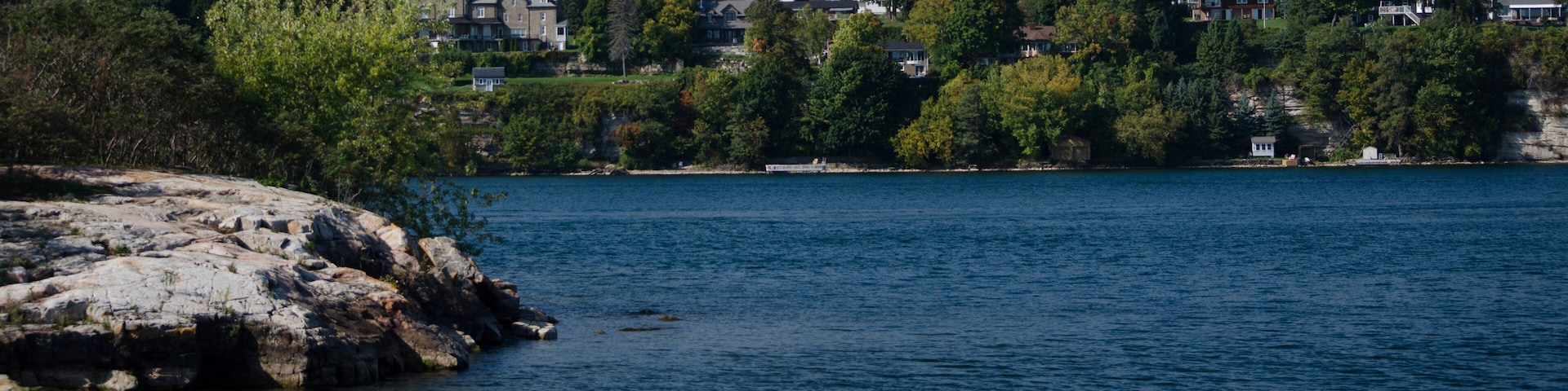A calm lake with rocky shores, surrounded by dense trees and houses on a hillside.