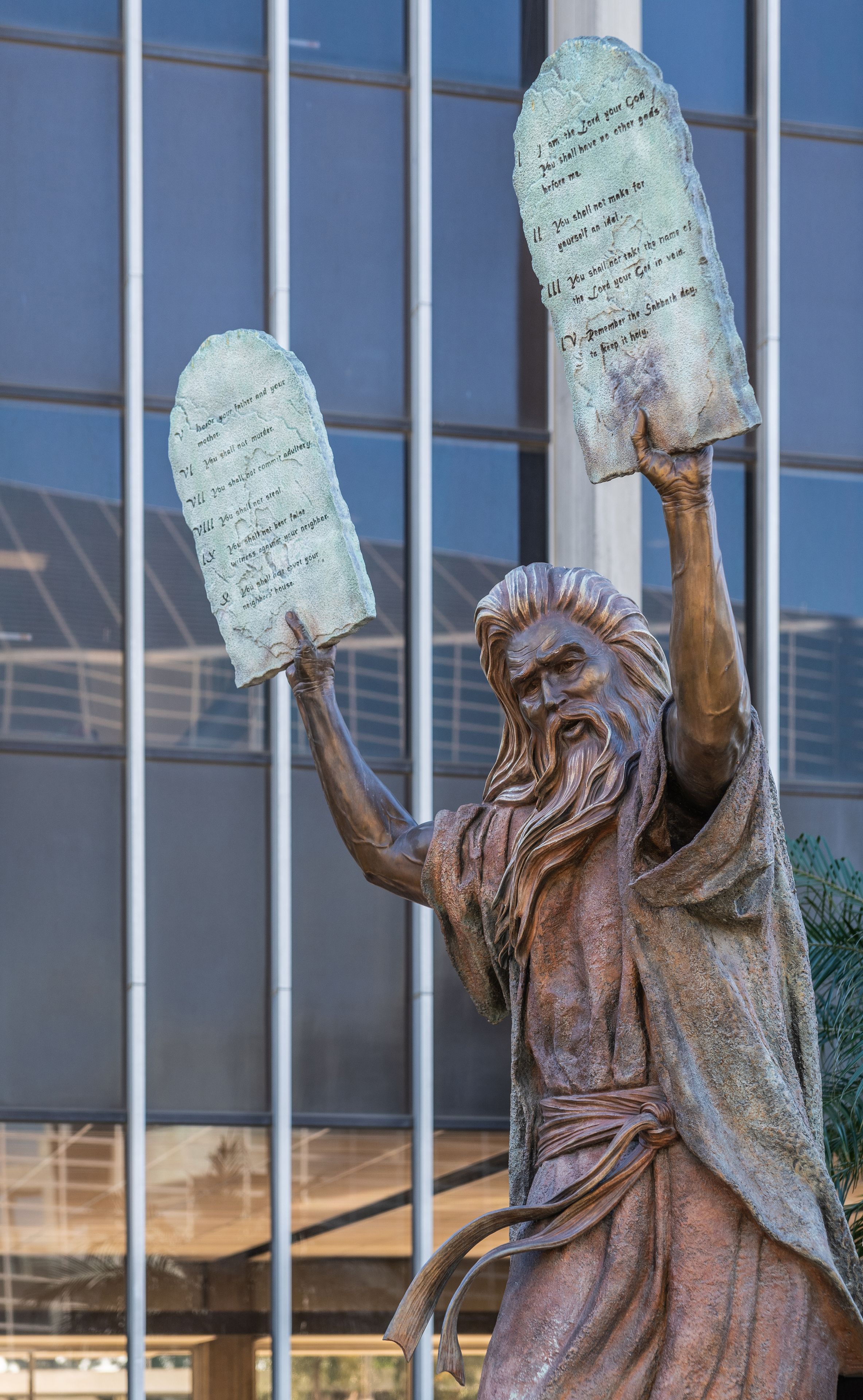 A bronze statue of a figure holding two stone tablets with inscriptions, set against a modern building facade.