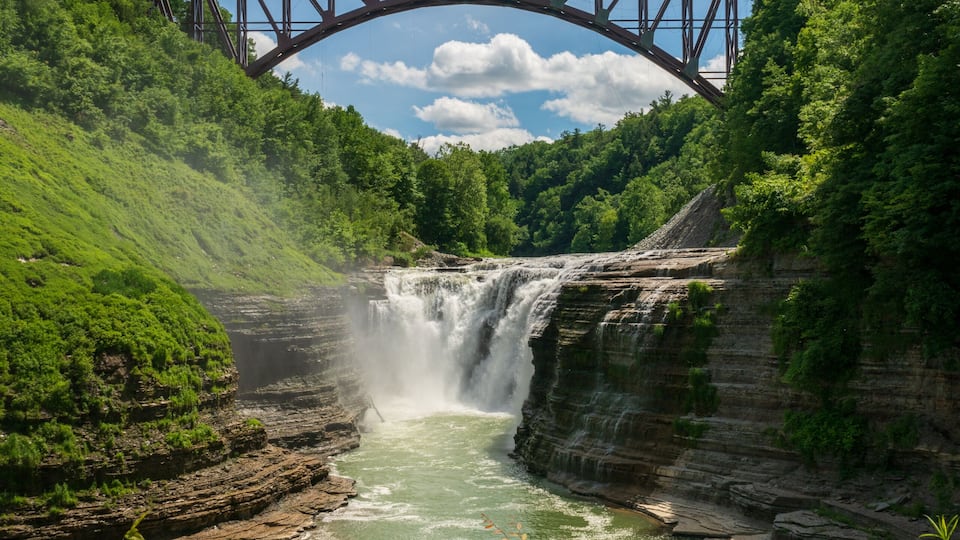 A waterfall cascading over a rocky cliff, a bridge spanning above, and lush greenery surrounding the area.