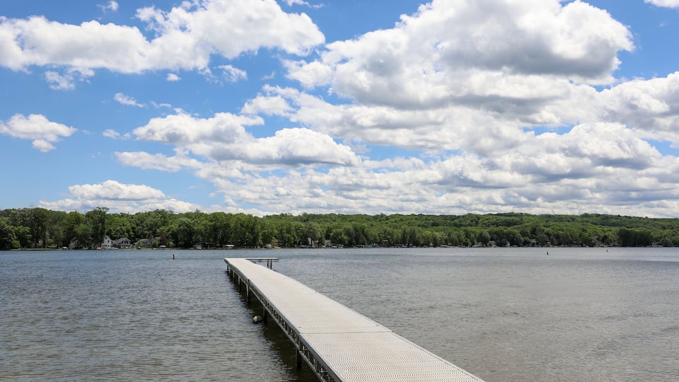 A long pier extending into a calm lake, surrounded by a forested shoreline under a partly cloudy sky.