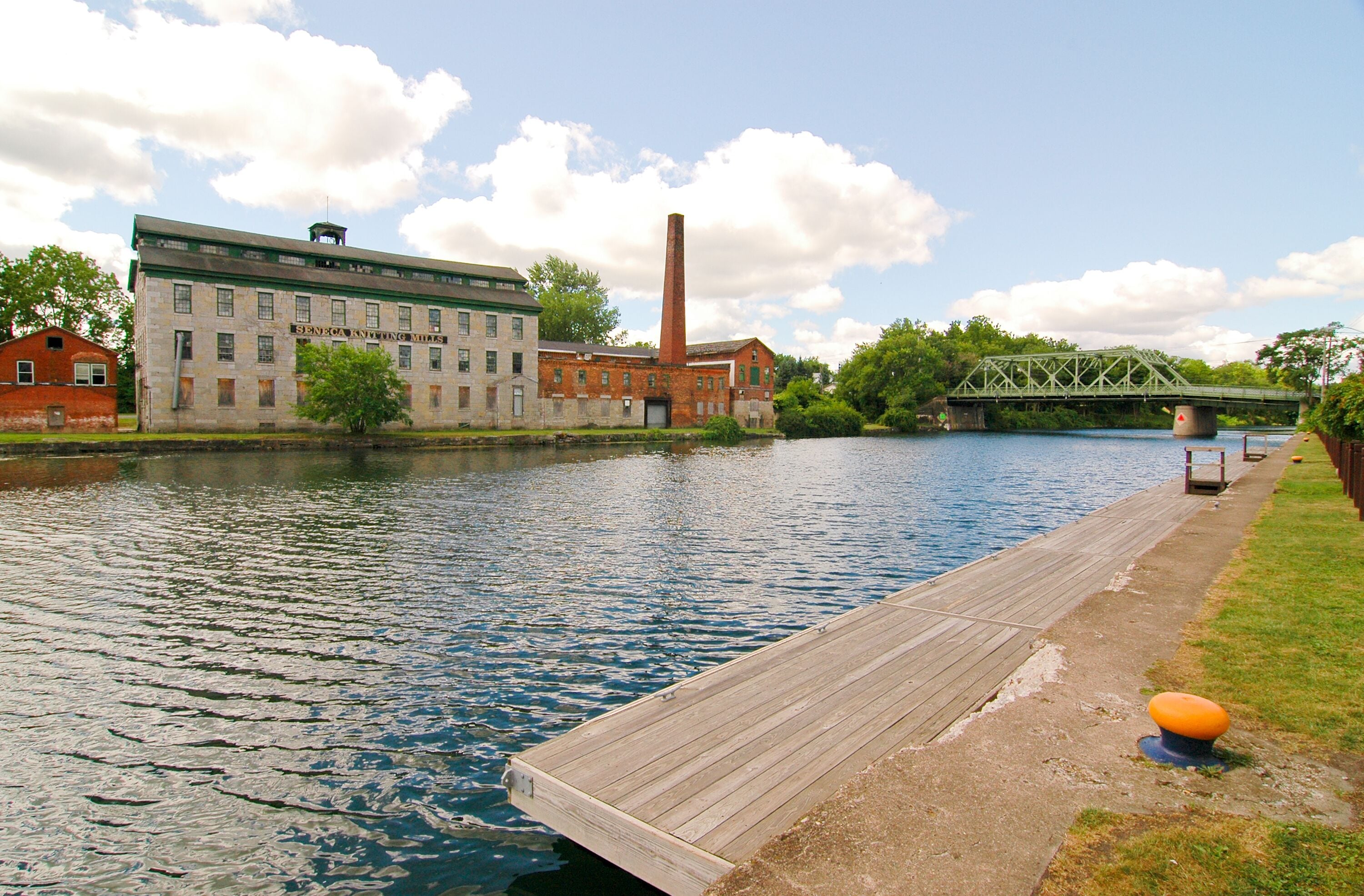 A river with a wooden dock, a brick building, and a green metal bridge.