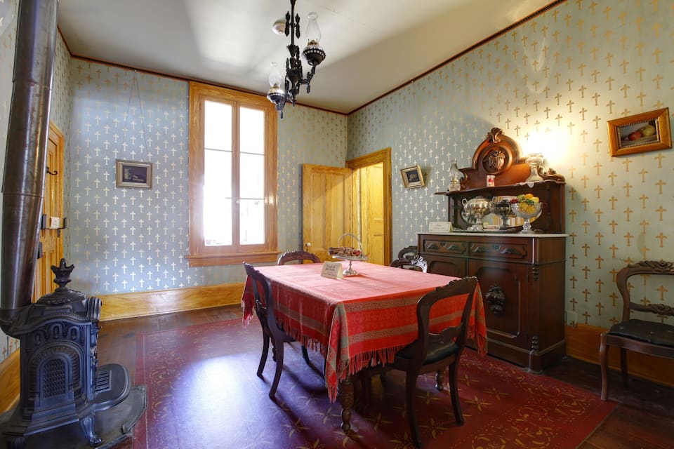 Uma sala de jantar com toalha de mesa vermelha, cadeiras de madeira, um lustre e papel de parede estampado.