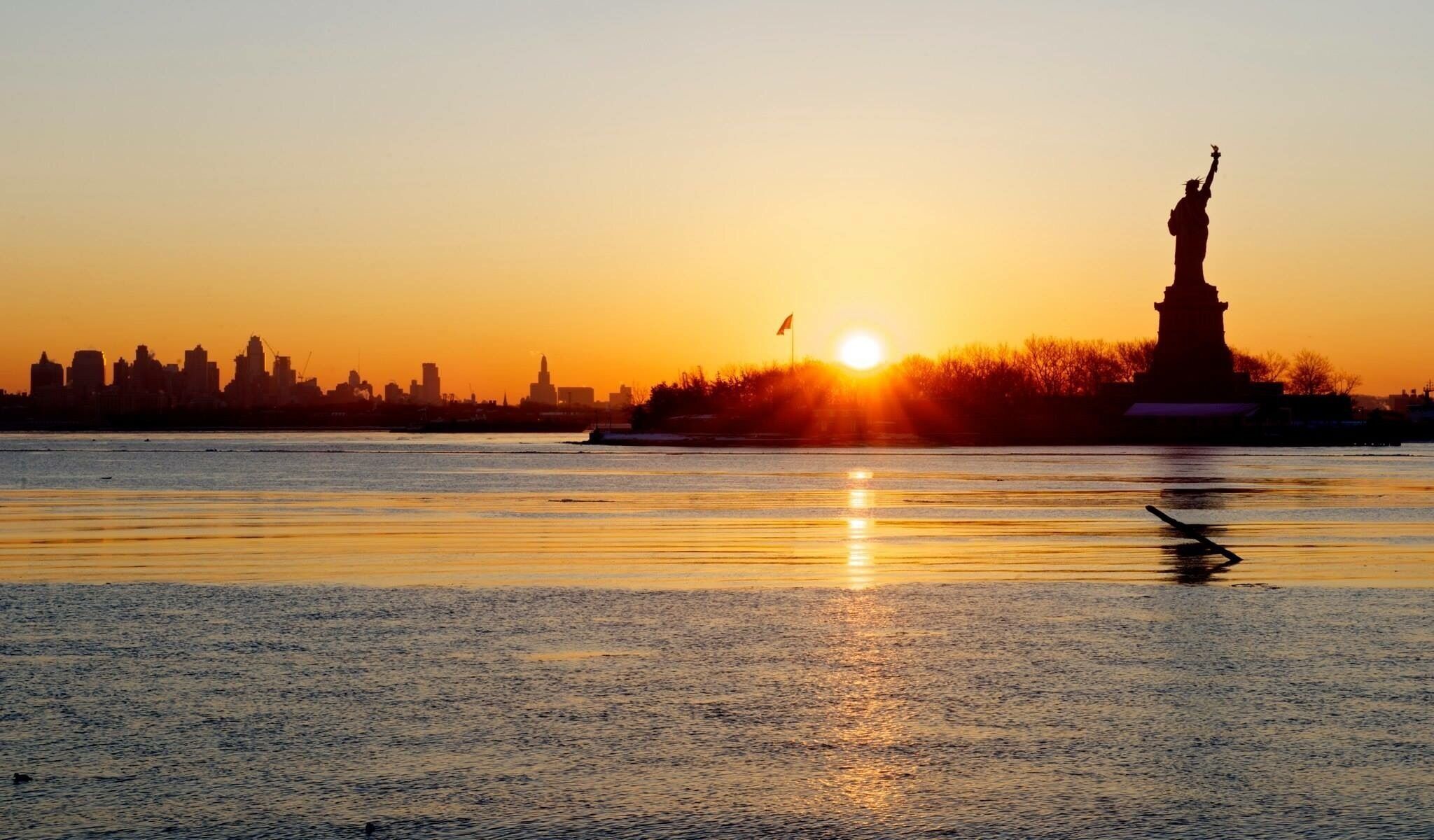 Een zonsondergang boven het water met een stadsachtergrond en een standbeeld aan de rechterkant.