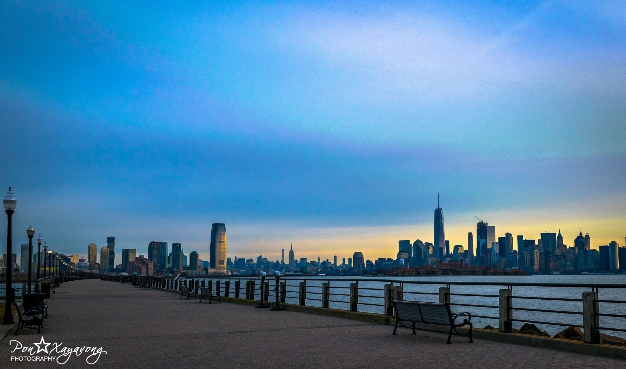 Un paseo marítimo con bancos, barandilla y el perfil del horizonte urbano al fondo.