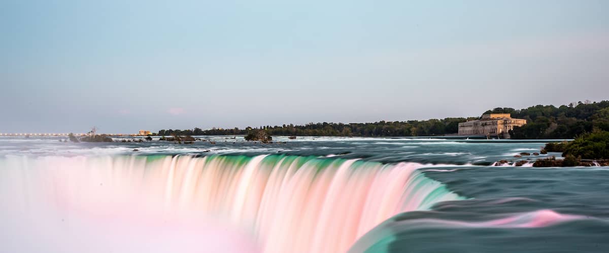 A waterfall with a pink and green hue, surrounded by a forest and a distant building.
