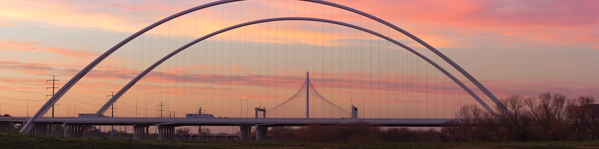A bridge with a distinctive arch design over a calm body of water, reflecting the structure and a sunset sky.