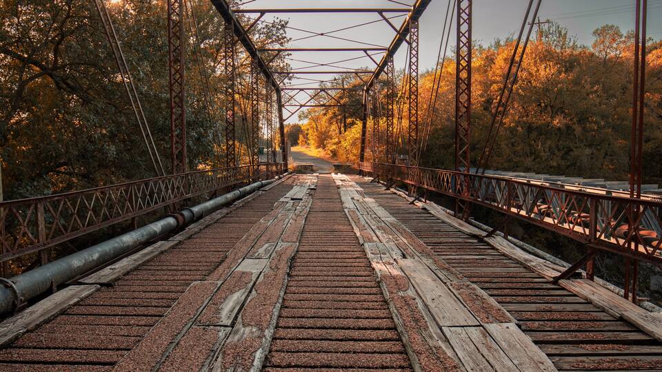 A bridge with a metal framework and wooden planks, surrounded by trees with autumn foliage.