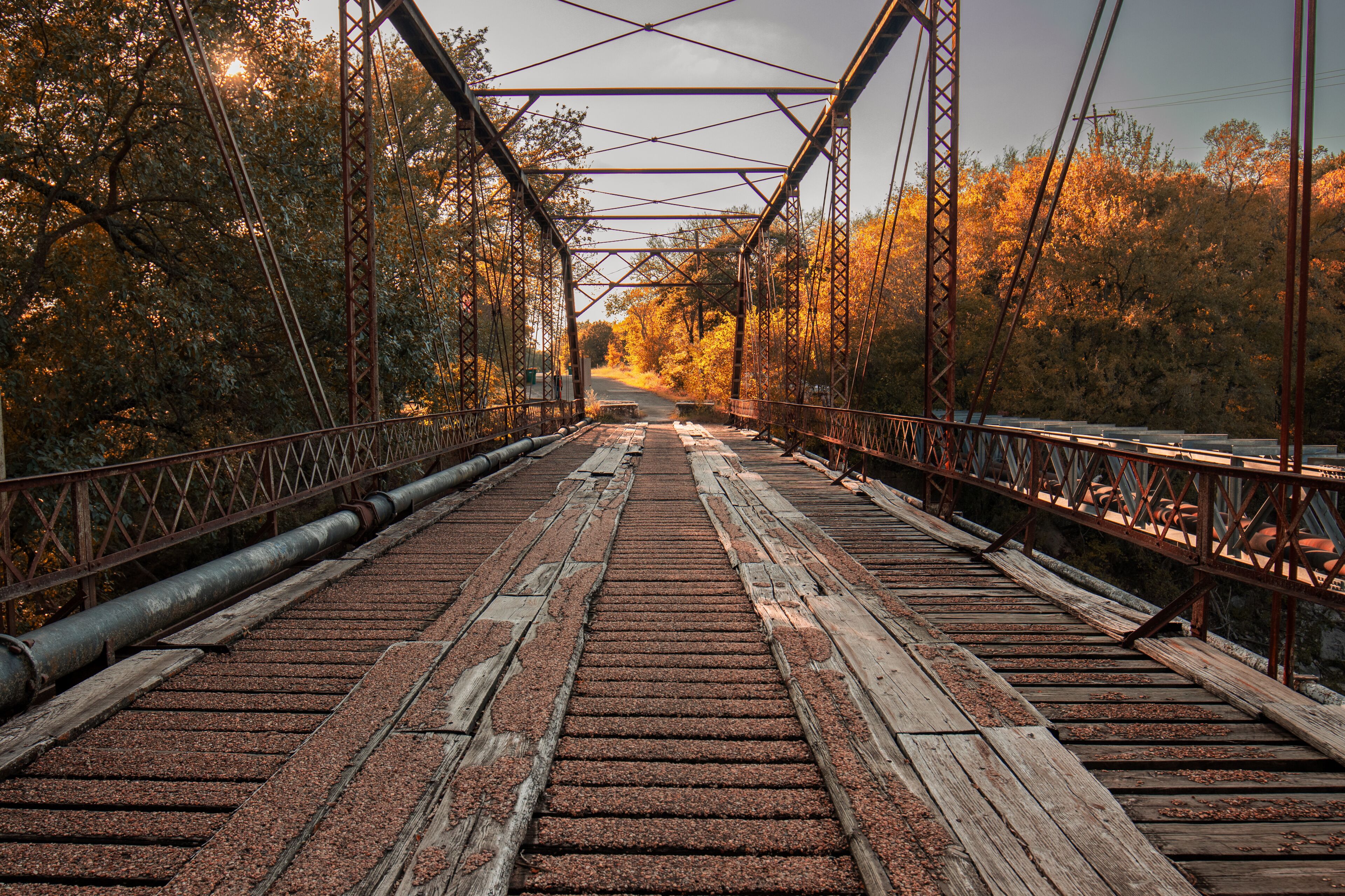 A bridge with a metal framework and wooden planks, surrounded by trees with autumn foliage.