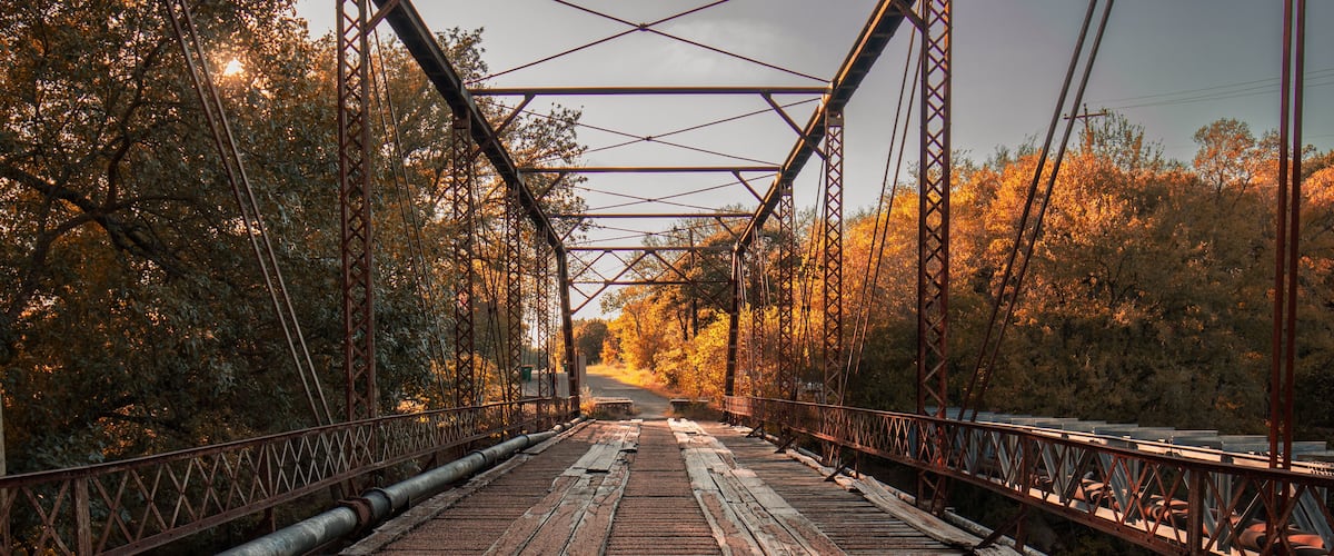 A bridge with a metal framework and wooden planks, surrounded by trees with autumn foliage.