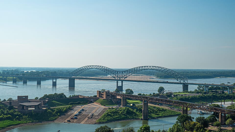 A large steel bridge spanning a wide river, surrounded by lush greenery and a parking area.