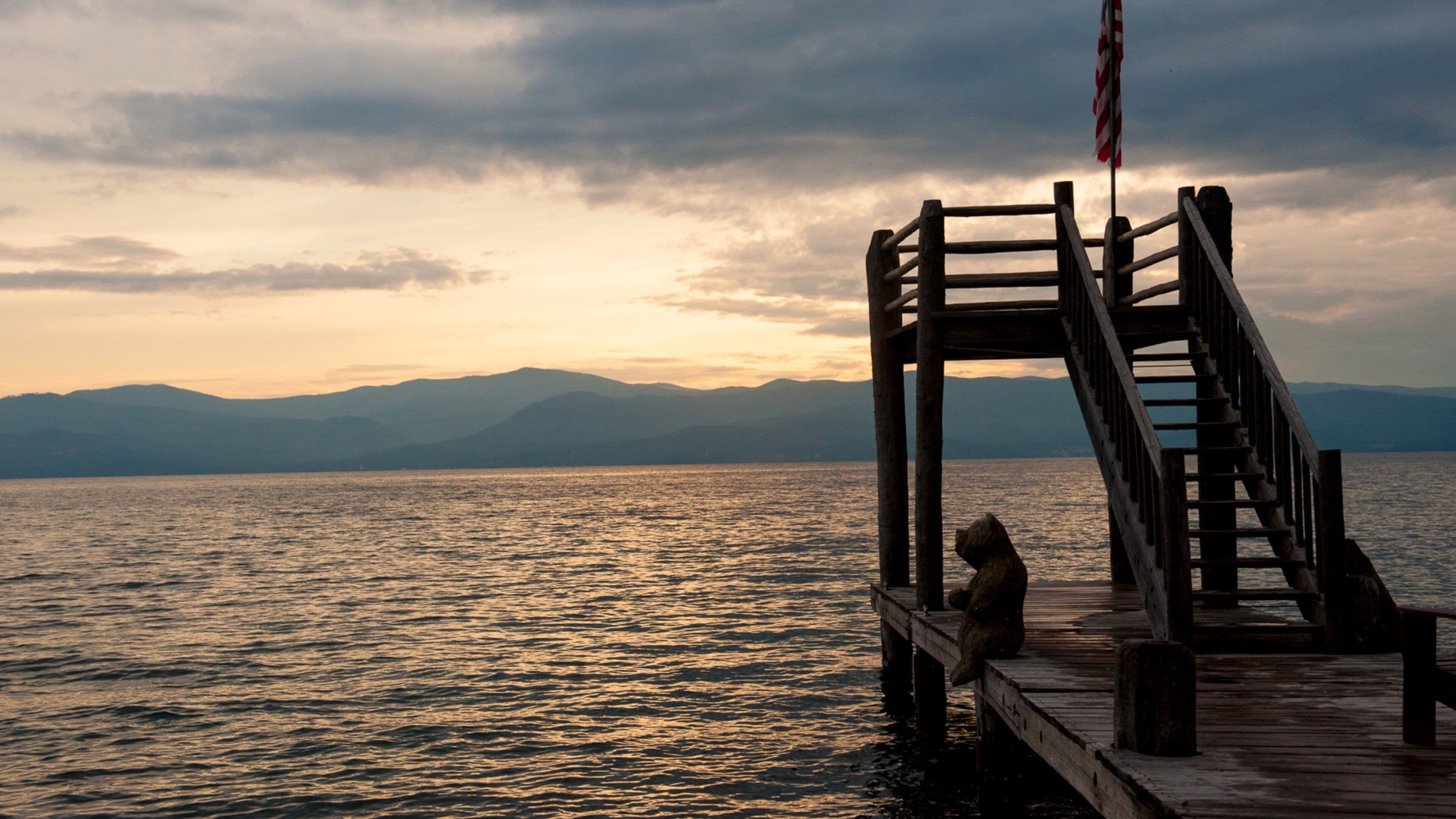 A wooden pier extending into a body of water with mountains in the background and an American flag on the right.