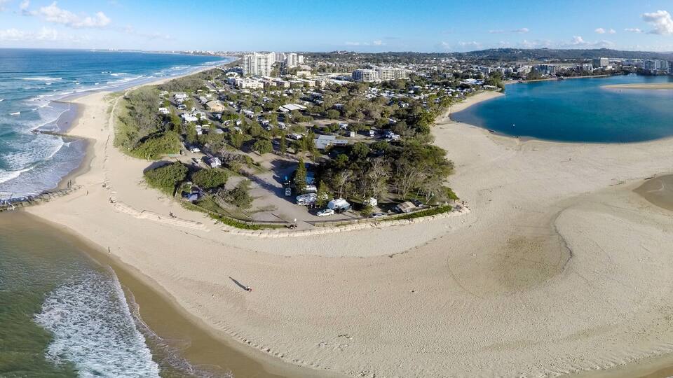An aerial view of a coastal area with a beach, a small island with buildings, and a clear blue sea.