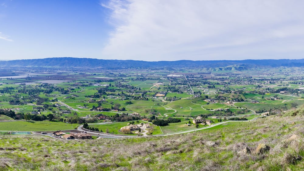 A panoramic view of a valley with rolling hills, a winding road, and a distant mountain range.