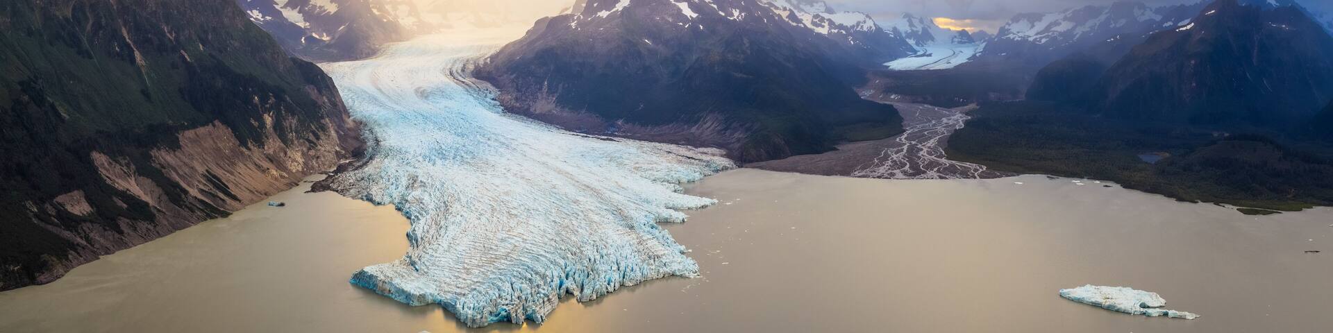 Aerial view of the majestic blue glacier flowing into a calm lake dotted with icebergs, cradled by rugged mountains under a dramatic sky, Cordova, Alaska, United States.