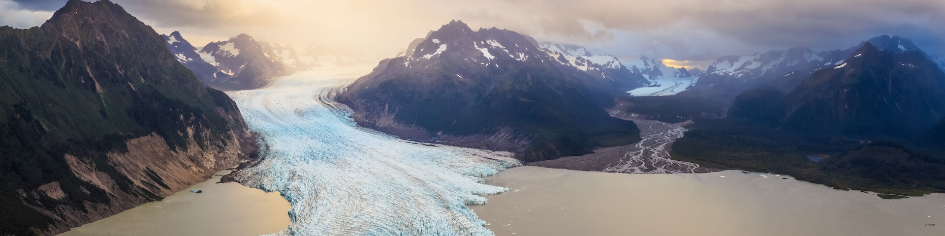Aerial view of the majestic blue glacier flowing into a calm lake dotted with icebergs, cradled by rugged mountains under a dramatic sky, Cordova, Alaska, United States.
