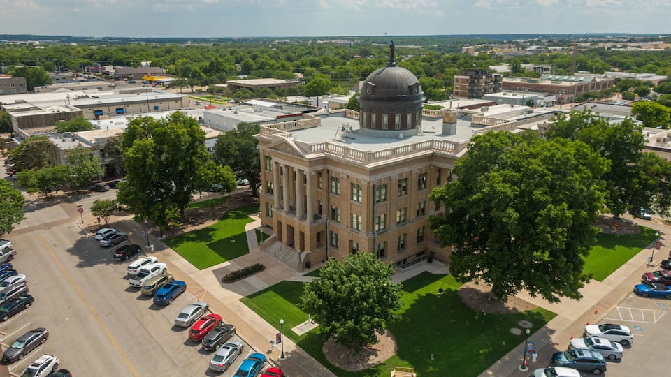 A large, historic building with a dome and columns, surrounded by a parking lot and greenery.