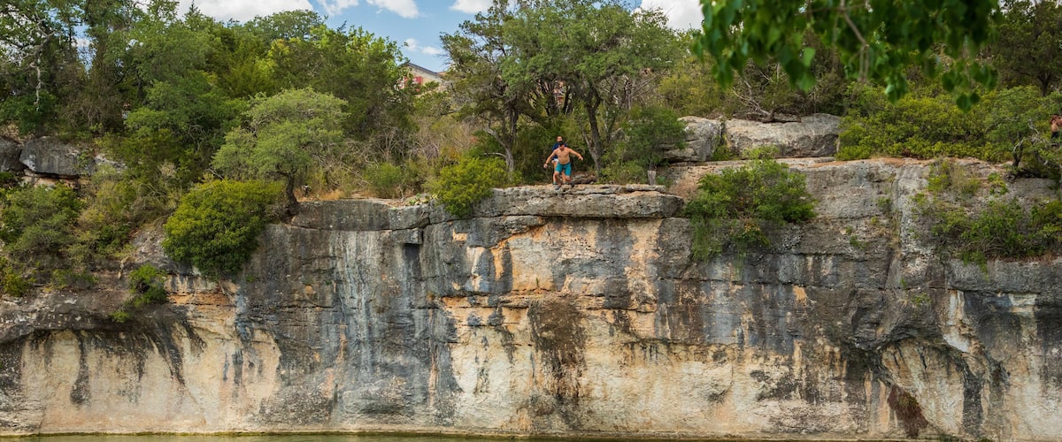 A rocky cliff with a person standing on top and another person swimming in the water below.