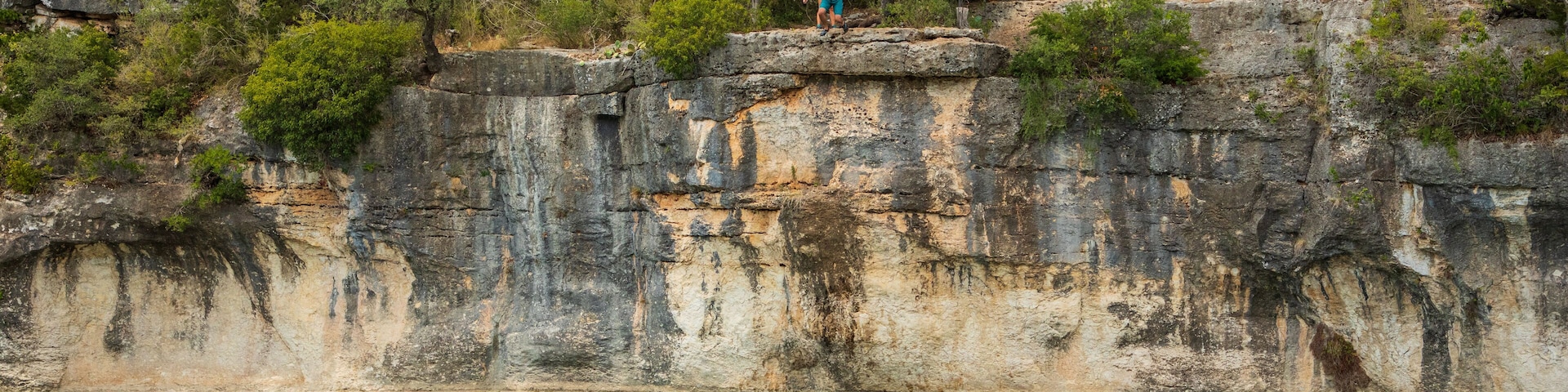 A rocky cliff with a person standing on top and another person swimming in the water below.