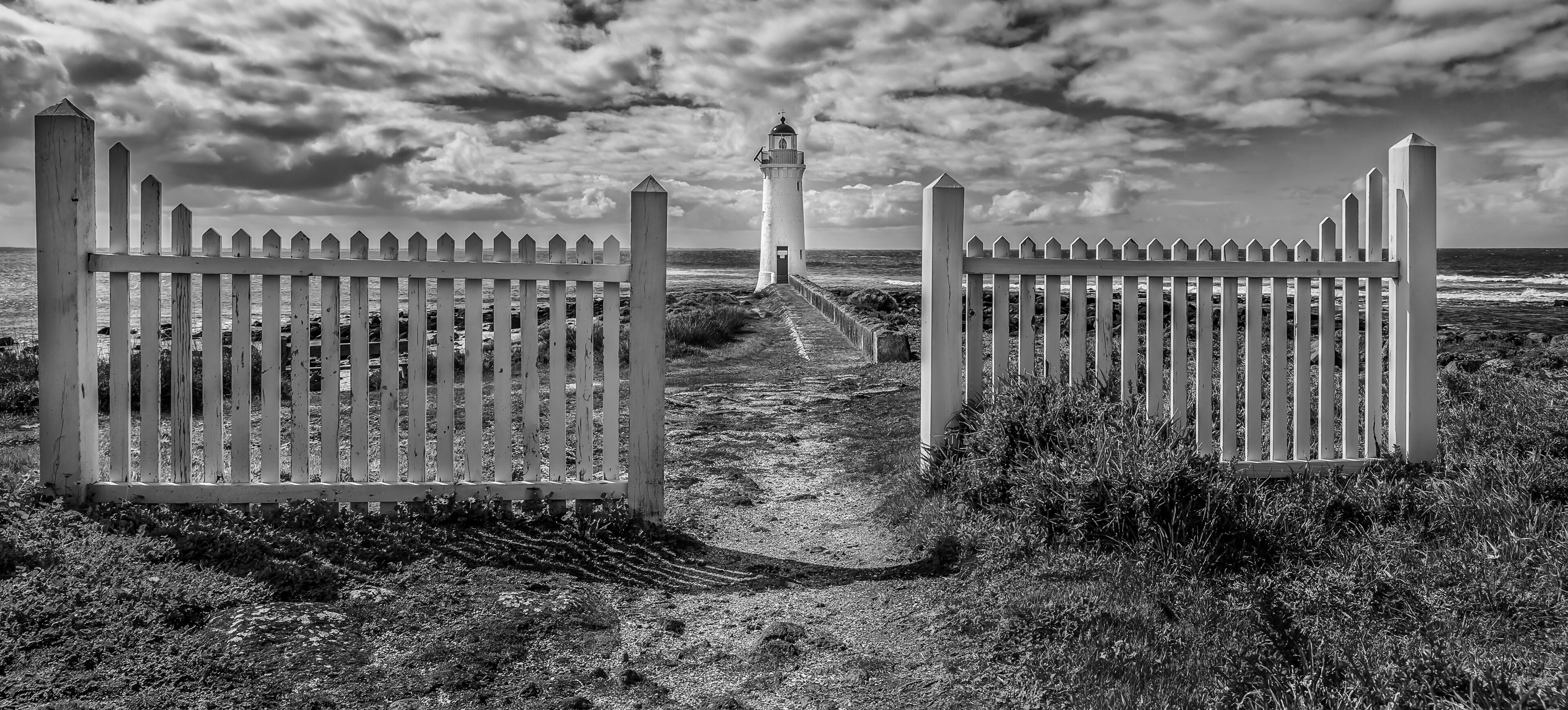 A coastal path leading to a lighthouse, flanked by white picket fences.