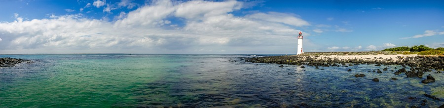 Un faro en una costa rocosa con aguas azules cristalinas y un cielo parcialmente nublado.