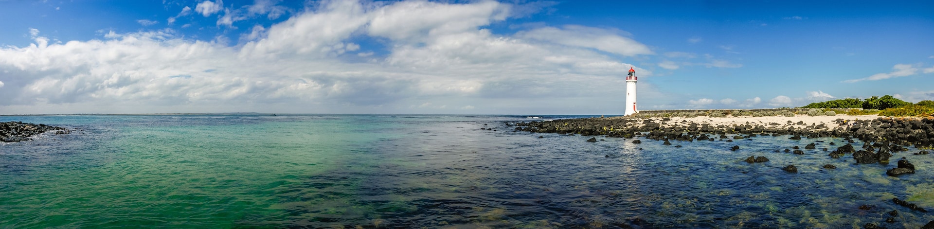 Un faro en una costa rocosa con aguas azules cristalinas y un cielo parcialmente nublado.
