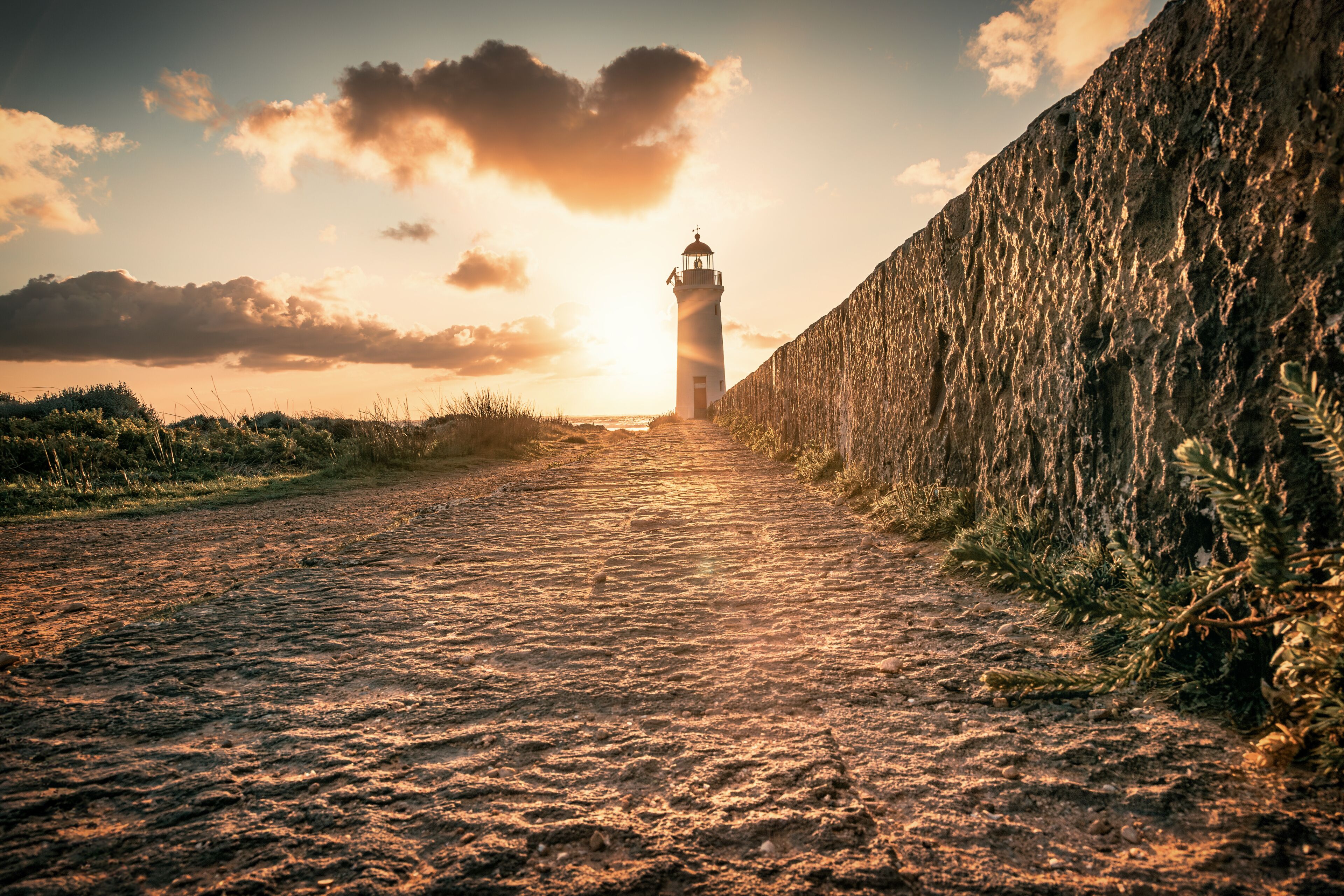 A coastal path leading to a lighthouse, with a rocky wall on one side and a clear sky with clouds.
