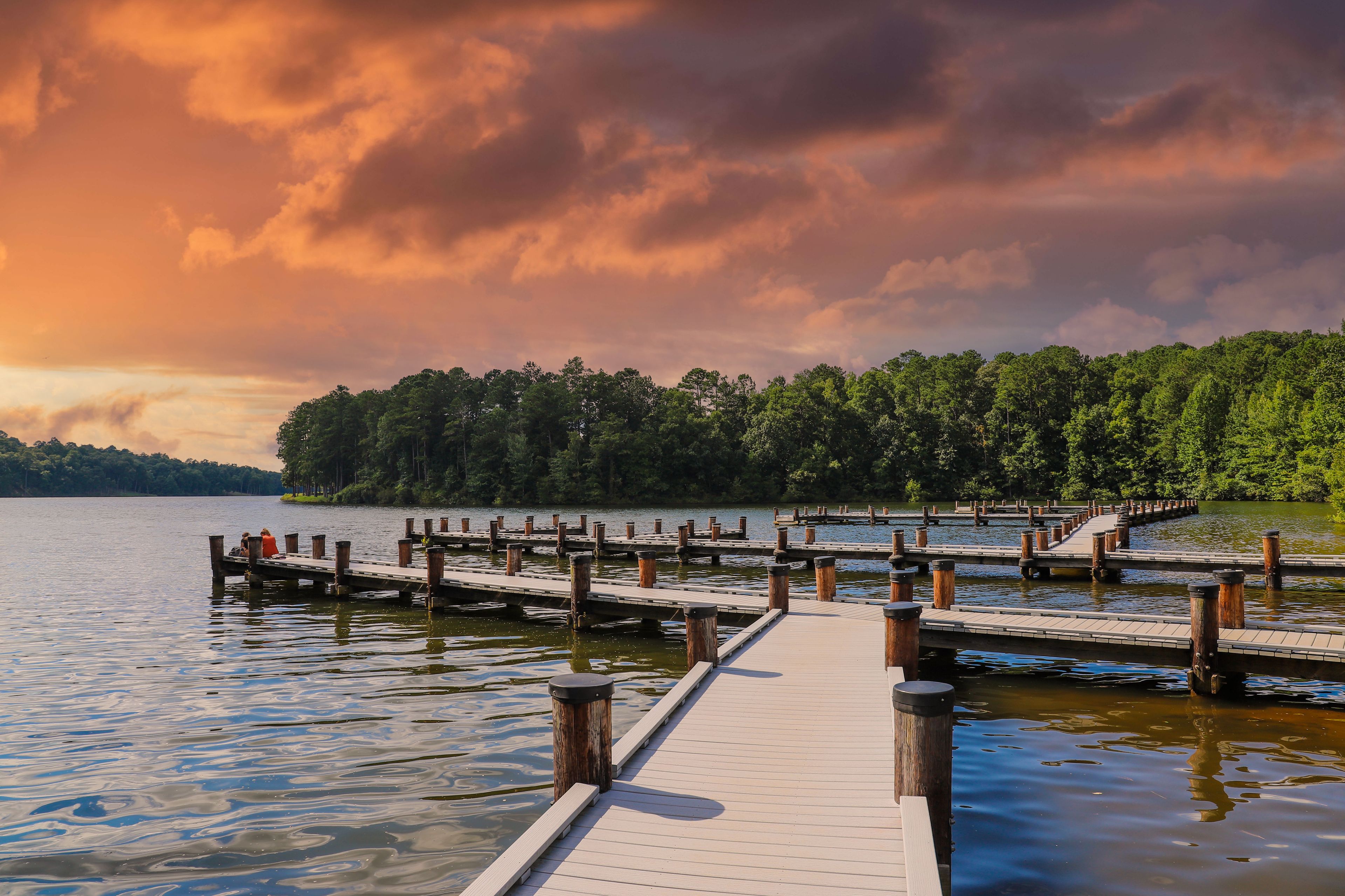 a gorgeous shot of the still blue lake water with a wooden dock over the water surrounded by lush green trees with powerful clouds at sunset at Lake Kedron in Peachtree City, Georgia USA