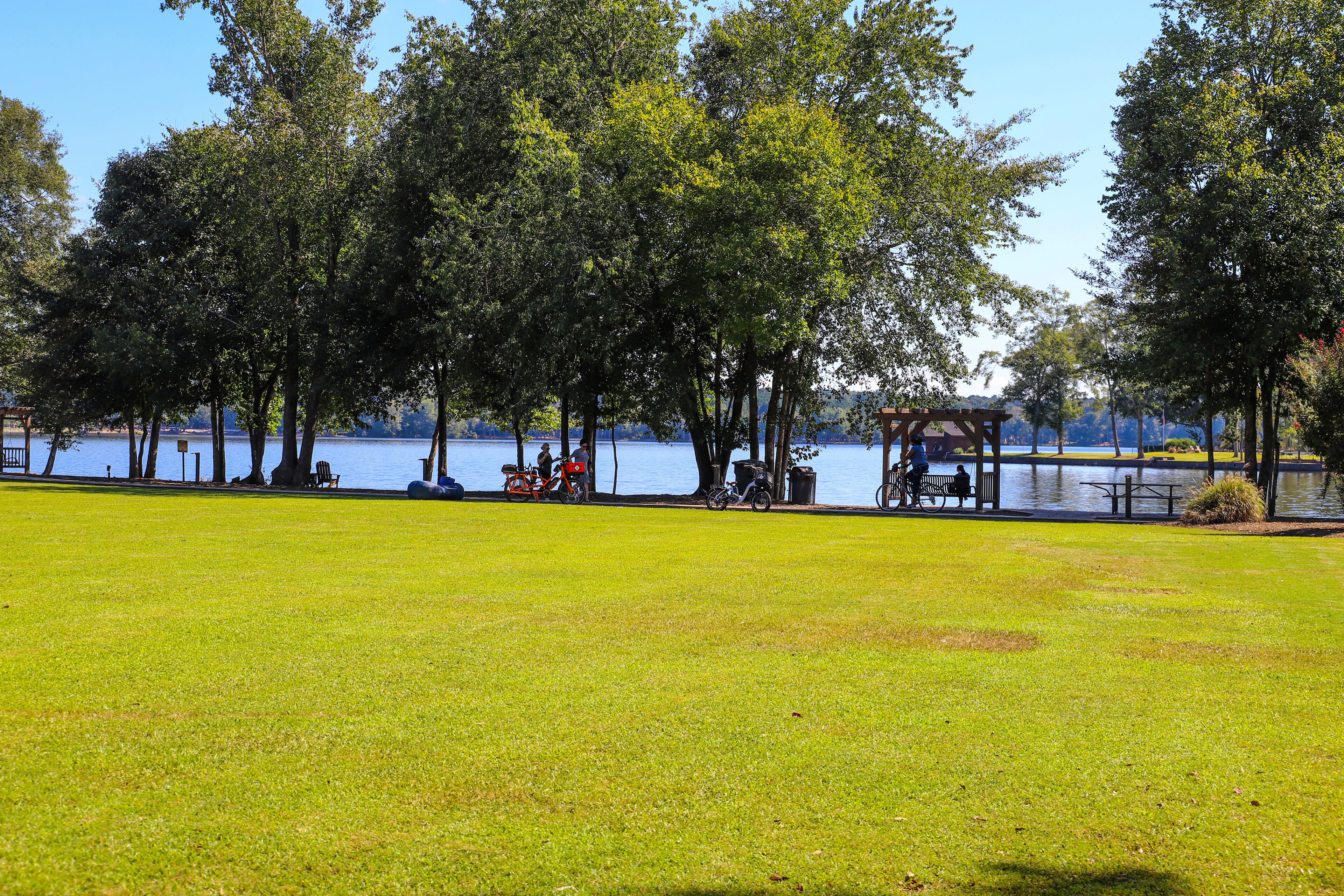 a shot of a park by the lake with vast green grass, lush green trees, people relaxing on the banks of the lake, blue sky and clouds at Lake Peachtree in Peachtree City Georgia USA