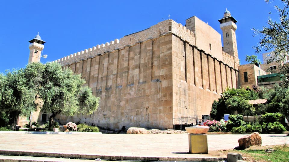 A historic stone building with two towers, surrounded by greenery and a paved area.