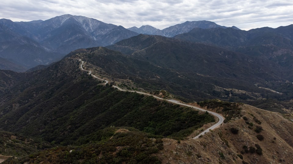 A winding mountain road through a mountainous landscape with dense vegetation and distant mountain peaks under a cloudy sky.