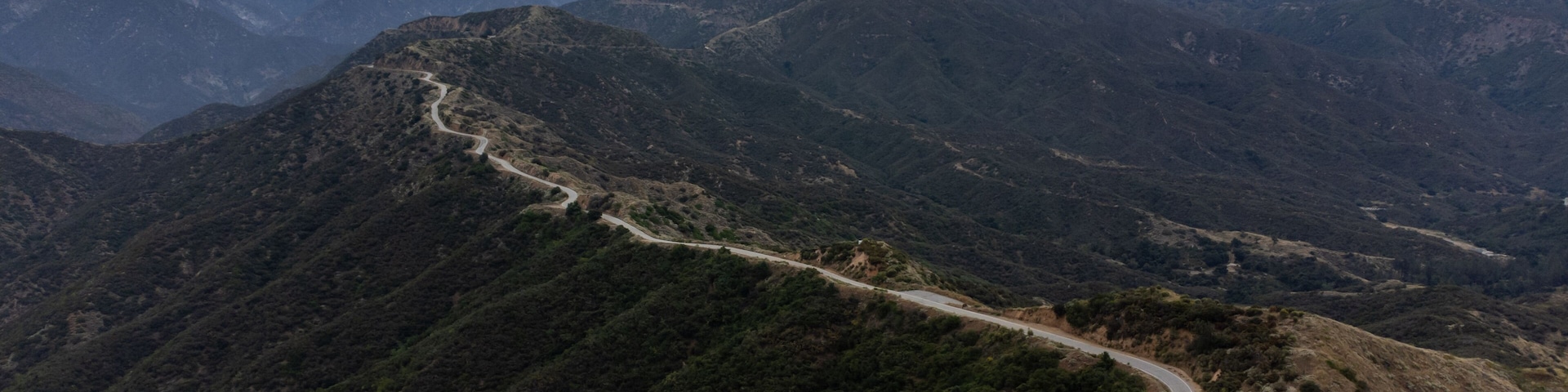A winding mountain road through a mountainous landscape with dense vegetation and distant mountain peaks under a cloudy sky.