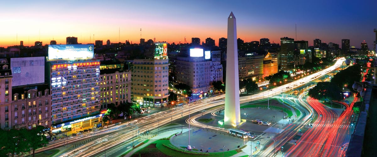 A cityscape at dusk with a prominent monument, illuminated buildings, and a busy intersection with light trails.