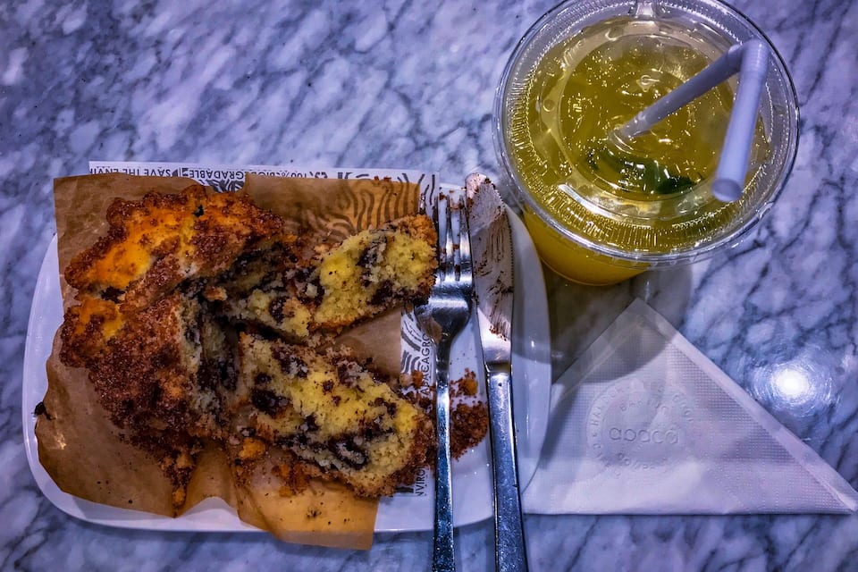 Een stukje taart met een kruimelige topping op een wit bord, een vork en een glas geel vocht met een rietje op een marmeren oppervlak.