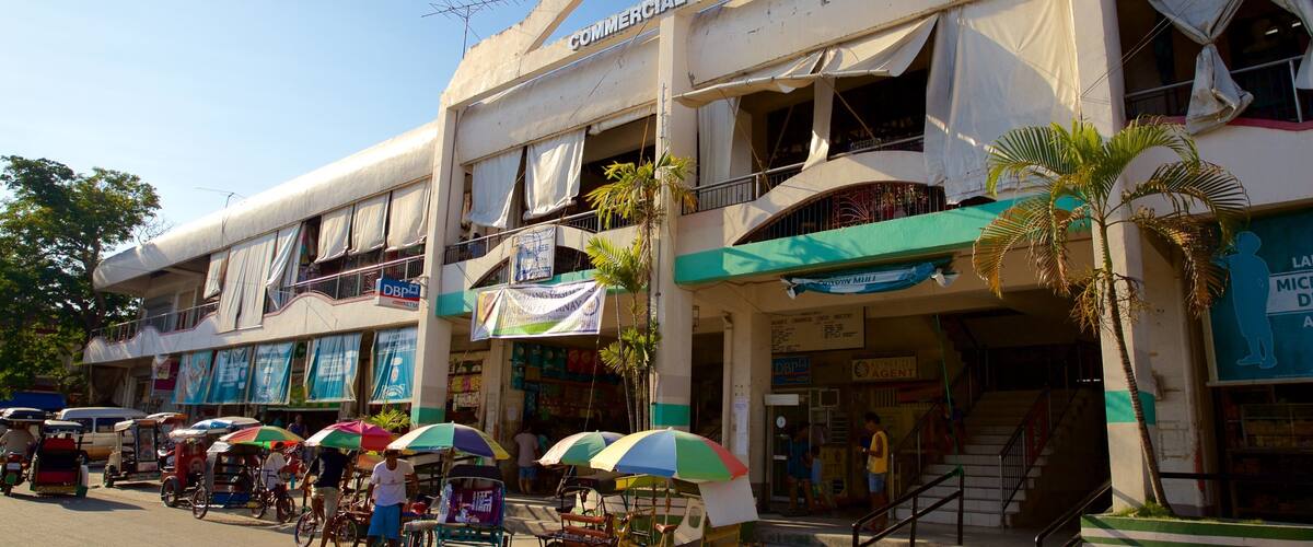 A commercial center with a sign reading "DALAGUETE COMMERCIAL CENTER". There are several shops with colorful umbrellas, a bicycle rickshaw, and a few people walking.