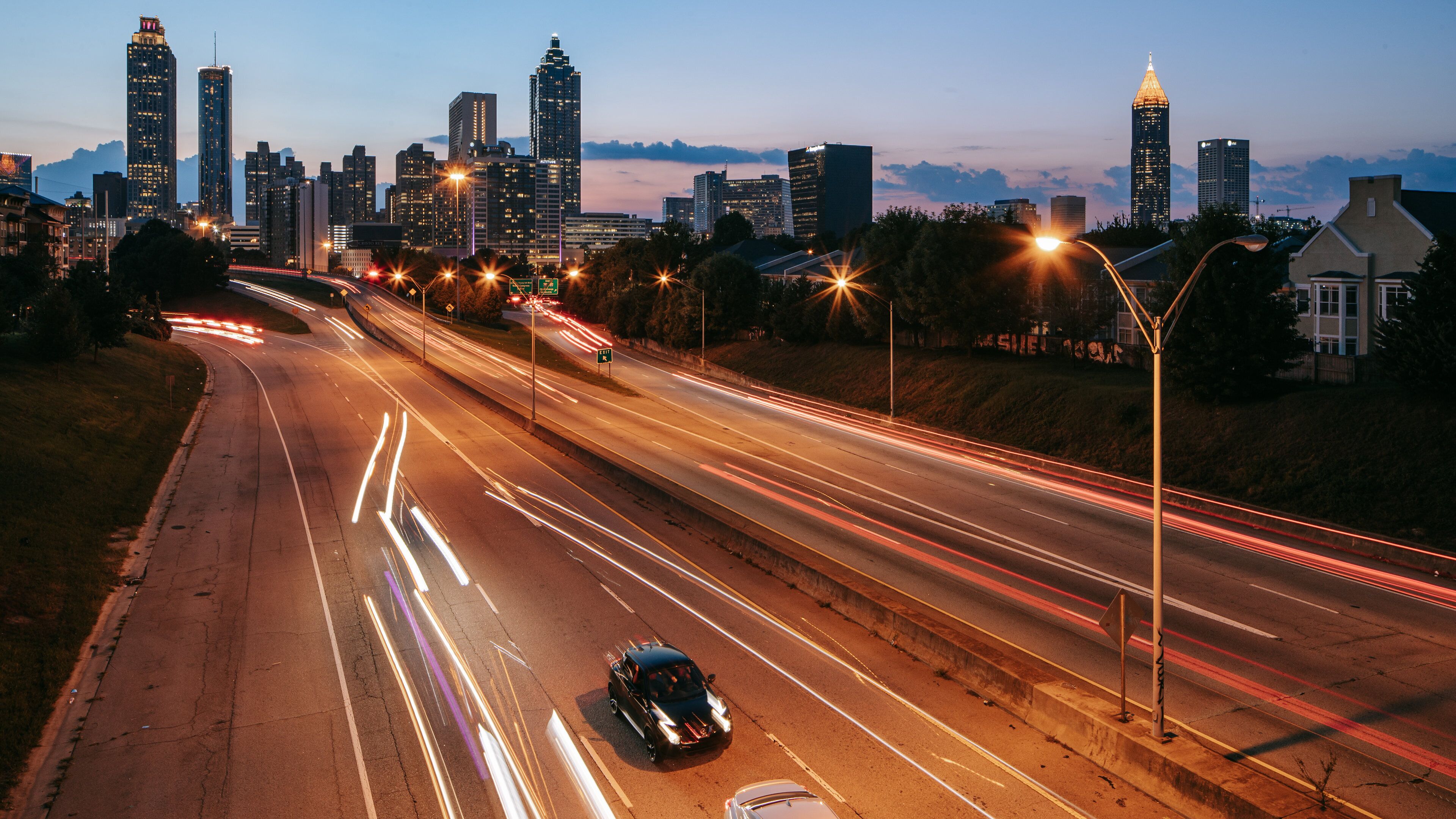 Ein Stadtbild in der Abenddämmerung mit beleuchteten Gebäuden und einer Autobahn mit Lichtspuren von vorbeifahrenden Fahrzeugen.
