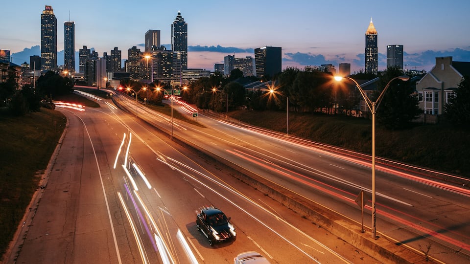 Ein Stadtbild in der Abenddämmerung mit beleuchteten Gebäuden und einer Autobahn mit Lichtspuren von vorbeifahrenden Fahrzeugen.