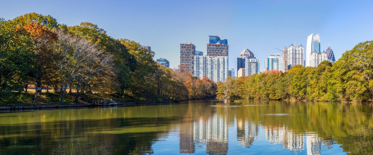 A calm lake reflecting trees and a city skyline with modern buildings.