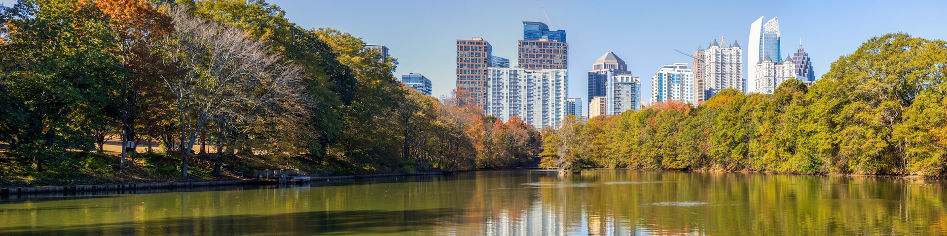 A calm lake reflecting trees and a city skyline with modern buildings.