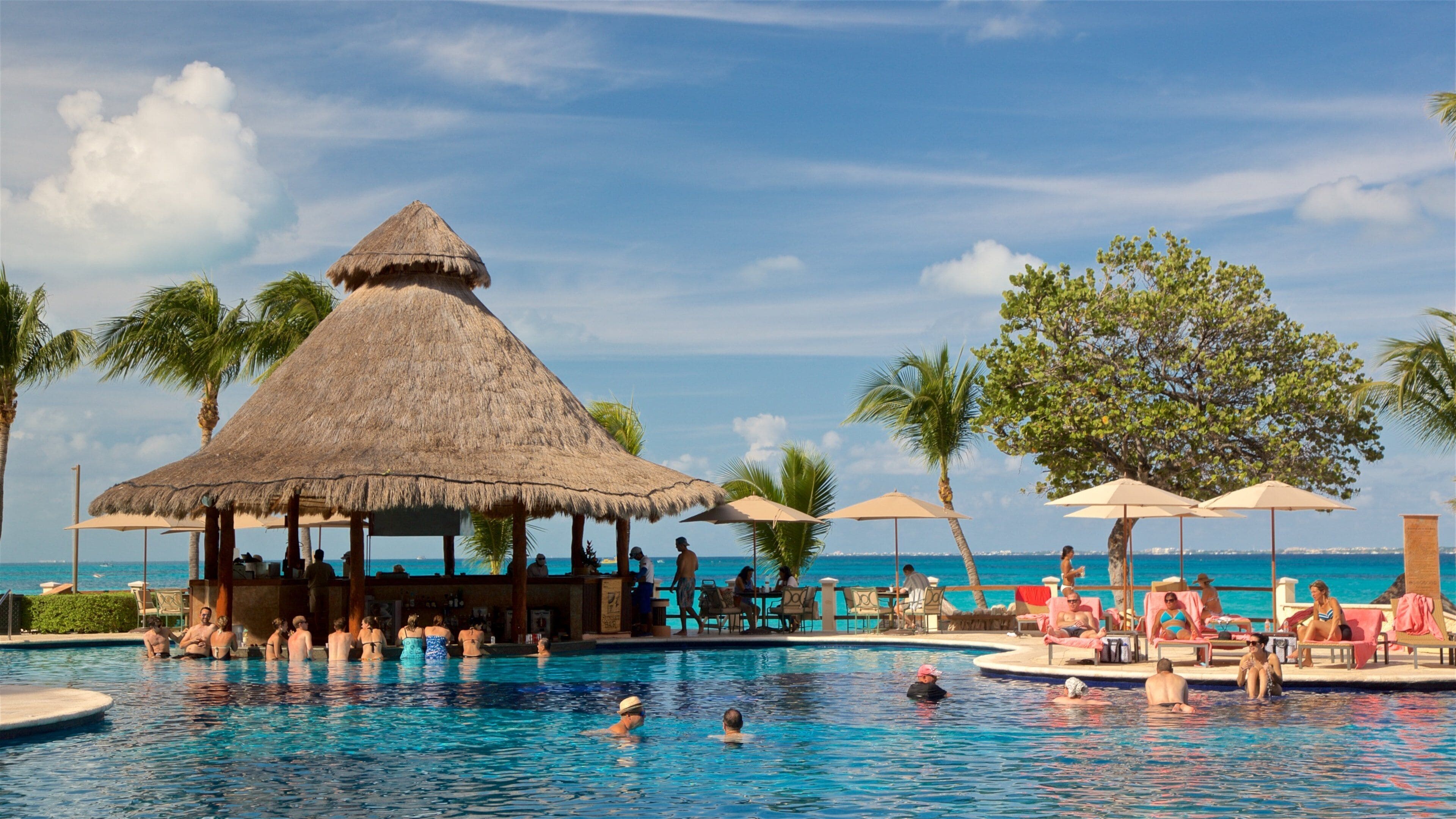 Ein Pool mit Schwimmern und Entspannungsbereich, umgeben von Palmen und einem Gebäude mit Reetdach.