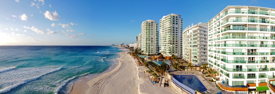 A beach with a clear blue ocean, adjacent high-rise buildings, and a pool area.