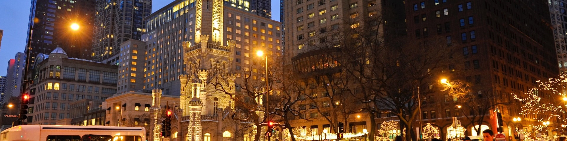 Rua urbana movimentada ao anoitecer, com edifícios altos, uma torre histórica e pedestres.