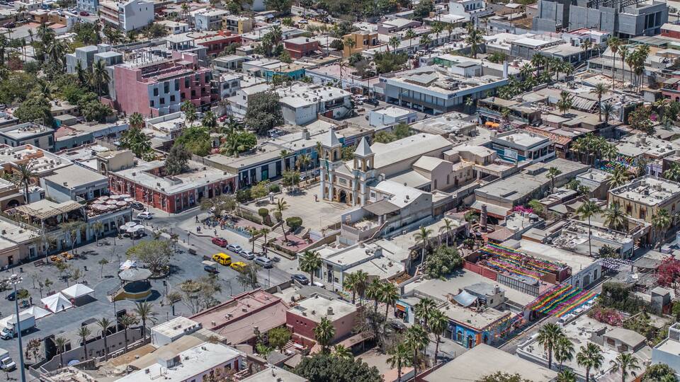 Une vue aérienne d’une zone urbaine densément construite, avec un mélange de bâtiments commerciaux et résidentiels, une église imposante dotée d’un clocher et une zone de marché aux couleurs vives.