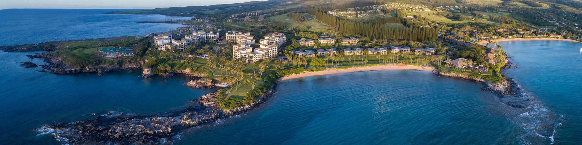 Vista aérea de um resort costeiro com praia, floresta densa e colinas suaves ao fundo.